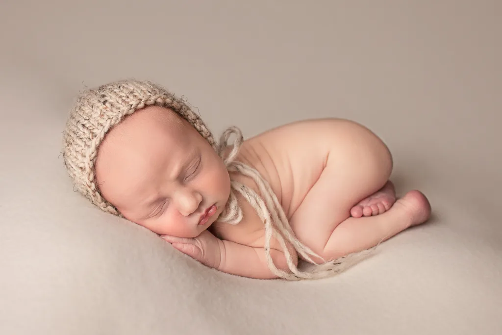 Image shows a newborn boy laying on a cream drop. He's wearing a beige knitted bonnet.